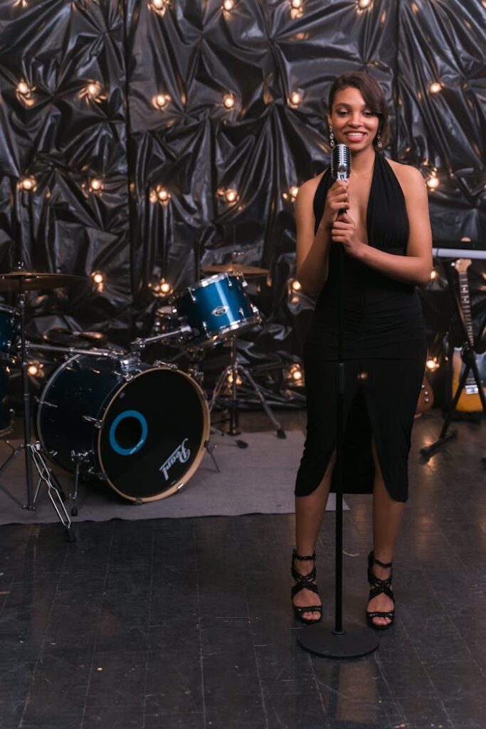 pexels-photo-8040854 African American woman in black dress singing on stage with a drum set and microphone.